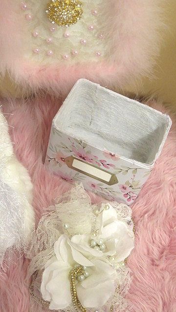 A white Victorian storage box with floral patterns and distressed details, displayed next to a pink furry surface, accompanied by a decorative floral wreath and a pair of pearl earrings.